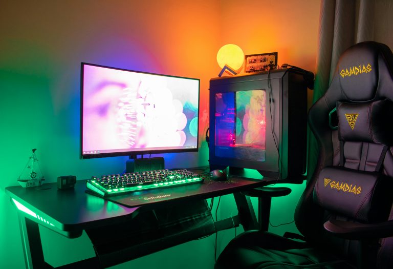 A colorful gaming setup featuring a PC, monitor, and gaming chair with RGB lights in a dark room.